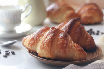 A cooked French croissant in a  plate with a cup of coffee in background. Sun light from window in...