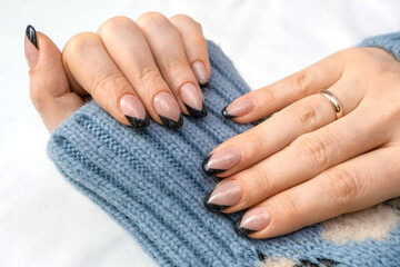 Beautiful French manicure with black contour and sequins. Well-groomed female hands with painted nails with gel polish on a cozy background of blue clothes. Fashionable color of gel nail polish.