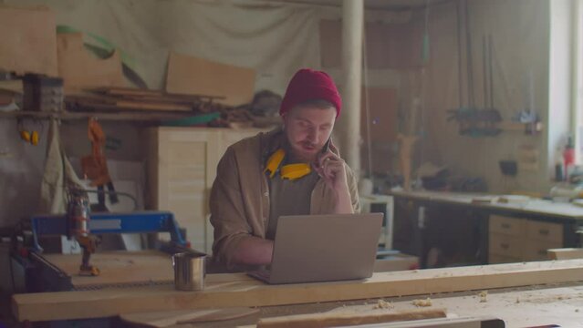 Caucasian male woodworker with safety earmuffs over his neck speaking on mobile phone and using laptop in carpentry workshop