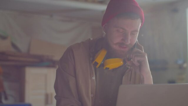 Professional woodworker with safety earmuffs over his neck talking on mobile phone and typing on laptop in carpentry workshop