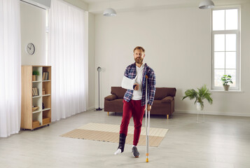 Physical injuries. Portrait of man who has injury to his arm and leg and wears special bandages and leans on crutch. Adult Caucasian bearded man in home clothes posing in living room looking at camera - obrazy, fototapety, plakaty
