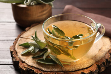 Cup of aromatic sage tea and fresh leaves on wooden stump, closeup