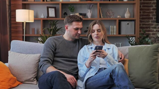 Couple family relaxing at sofa using smart mobile phone for online shopping. Happy millennial couple using funny apps smiling man and woman relaxing at home, having fun on social media online.