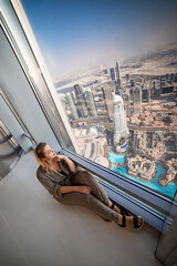 Beautiful tourist girl sitting by the window in Dubai Burj Khalifa tower with an amazing panoramic view over the city and fountains.