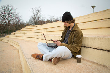 young man in cap wearing technological devices sitting on stairs outdoors