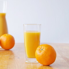 There is a glass of freshly squeezed orange juice on a wooden table, next to it lies a whole orange. A jug of juice and an orange can be seen in the distance.