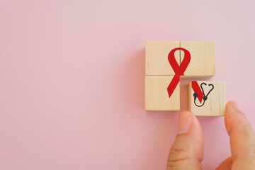 senior's hand complete red ribbon icon any stethoscope on wooden cube block with pink background for World Aids Day, world cancer day concept
