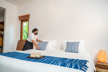 Asian hotel maid setting up pillow on bed sheet in hotel room.