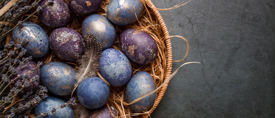 Obraz premium Purple, blue and golden eggs in a basket on a dark background. The purple hue trend of 2022 is very peri. Natural dye karkade tea. Top view. Easter card with a copy of the place for the text.
