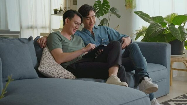 Young smiling gay couple using smartphone and relaxing in the living room at home, LGBTQ and diversity concept.