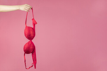 A woman's hand holding a red bra on a pink background. The concept of women's breasts, underwear. Copy space - obrazy, fototapety, plakaty