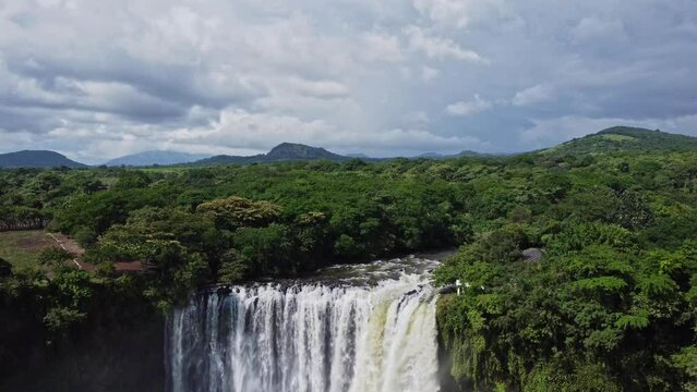 Tilt down shot featuring a beautiful waterfall in the Mexican jungle is surrounded by lush green vegetation on both sides with the view of hills in the background.