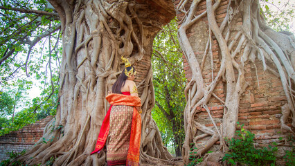 Asian woman in a Thai traditional dress walk to an old brick gate with a big root tree cover wall