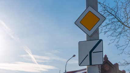 Road sign main road in sun beams against blue sky with clouds. Priority road sign on the background of sky and old building.