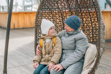 grandmother and granddaughter sat down to rest after a walk in a hanging rattan chair outdoors. an adult woman and a child are resting after a walk in autumn, spring on the street in outerwear