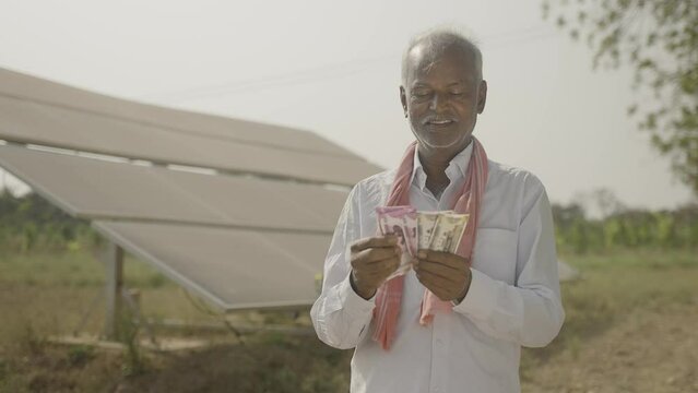 happy smiling indian farmer couting money while standing in front of solar panel at farmland - concept of electric power savings, investment, banking and financial