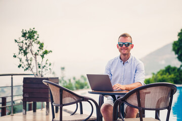 Successful handsome male businessman in sunglasses works at a laptop sitting near the pool. Remote work. Freelancer