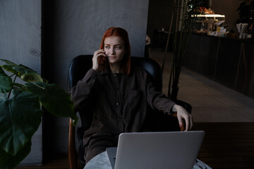 a red-haired freelance woman with glasses is talking on the phone, working remotely in a cafe. there is a laptop nearby.