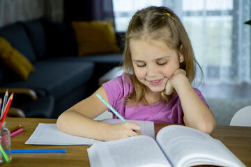 cute little girl blonde does school lessons sitting at a table in the living room. He writes in pencil in a notebook and smiles. The concept of homeschooling, education.