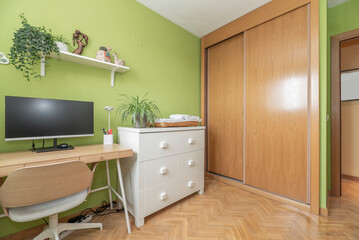 Desk with pc on wooden table, chest of drawers with six drawers and built-in wardrobe with sliding oak doors in a room with pistachio green walls