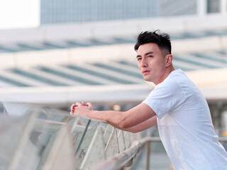 Portrait of handsome Chinese young man in white T-shirt standing and looking away with modern city buildings background in sunny day, side view of confident young man.