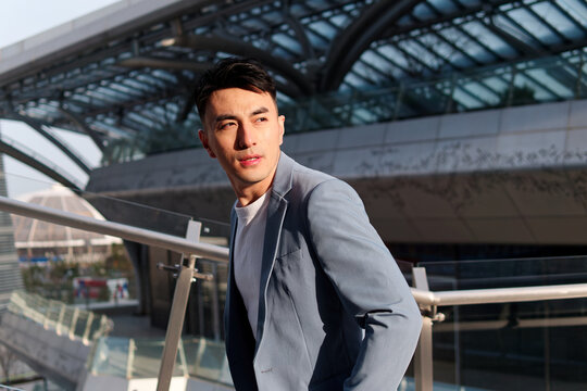 Portrait of handsome Chinese young man in light blue suit and white undershirt walking and looking away with modern city buildings background in sunny day, side view of confident businessman.