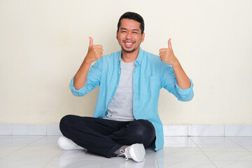Full body portrait of adult Asian man sitting cross legged smiling happy and give two thumbs up