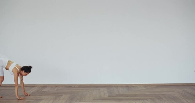 Flexible young woman in white sport bar and leggings doing wheel pose indoors. Caucasian female spending free time for regular workout at gym.