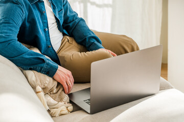 A young man in a blue shirt is working with a laptop while sitting on the couch at home in a cozy room. The concept of using the technology of the contact communication device business, IT technolog