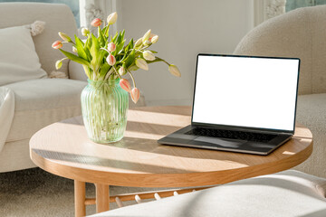 A laptop with a blank screen for your text on a wooden table in a modern bright interior with a bouquet of spring flowers