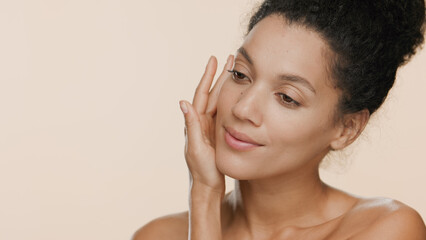 Young slim pretty black-haired African American woman with naked shoulders strokes her face with palm of her hand against beige seamless background | Smooth face concept