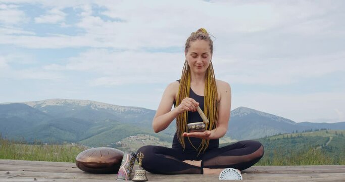 Happy young woman with braided hairstyle sitting in lotus pose and playing on tibetan singing bowl. Female hipster spending leisure time for sound therapy on fresh air.
