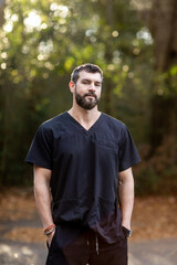 A doctor with dark hair and a beard in black scrubs standing outside in a natural green environment