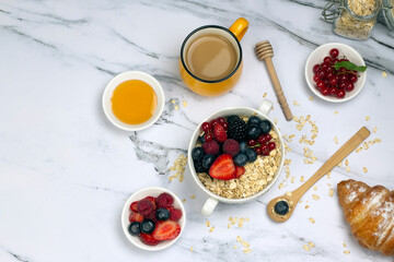 Healthy breakfast with cup of coffee, oat with berry, honey, croissant and ripe sweet berries on white marble table. top wave, flat lay.