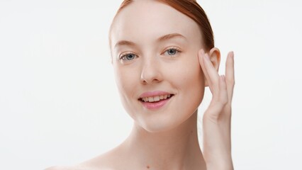 Obraz na płótnie Canvas Young positive ginger woman touches her cheek with her finger against light-grey background | Skin care commercial concept