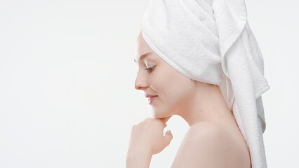 Cute slim European young woman with a towel on her hair touches her chin standing sideways to the camera on white background | Skin rejuvenation concept