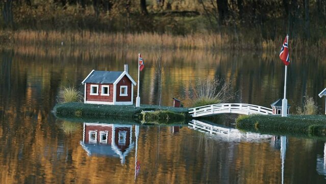 An idyllic scene in the town park in Finnsnes, Norway. Toy houses in the middle of the lake reflected in the mirrorlike still water. Colorful autumn foliage in the background. Slow-motion, pan left