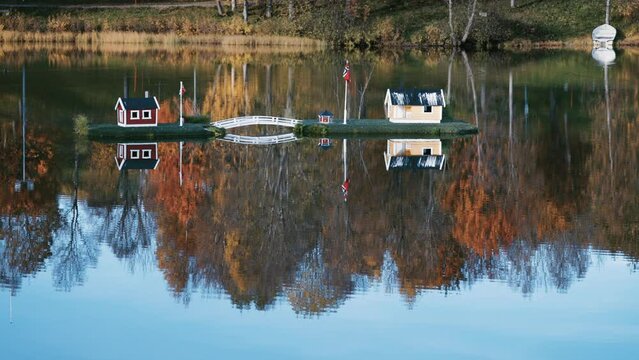 An idyllic scene in the town park in Finnsnes, Norway. Toy houses in the middle of the lake reflected in the mirrorlike still water. Colorful autumn foliage in the background. Slow-motion, pan right