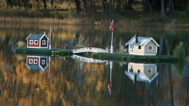 An idyllic view in the town park in Finnsnes, Norway. Toy houses in the middle of the lake reflected in the mirrorlike still water. Colorful autumn foliage in the background. Slow-motion, pan left