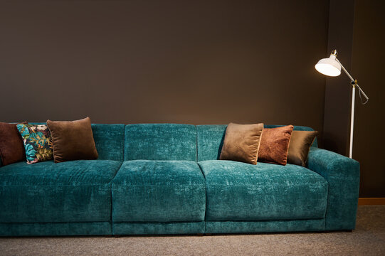 Exhibition of modern stylish upholstered furniture in the showroom of a furniture store. Focus on a turquoise soft velour setee and brown pillows lit by a lamp against a brown wall background