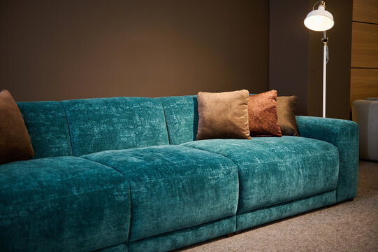 Exhibition of modern stylish upholstered furniture in the showroom of a furniture store. Focus on a turquoise soft velour setee and brown pillows lit by a lamp against a brown wall background