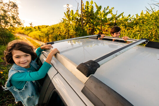 Crianças sorrindo sentadas na janela de um carro numa estrada de terra.