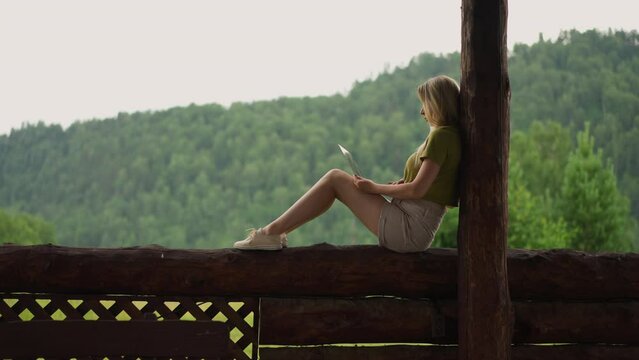 Concentrated woman programmer works on modern laptop sitting on rustic porch at eco resort at highland at drizzling rain side view slow motion