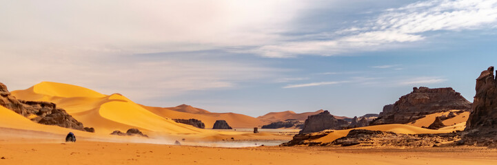 Fototapeta premium Panoramic view of Sahara Desert sand dune and rocky mountain off road nature.Tadrart Rouge, Djanet, Illizi. Erg dusty road. Orange colored sandstones and white sand powder in the air. Blue cloudy sky.