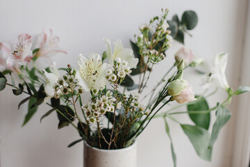 Stylish bouquet in ceramic vase at window, moody image. Beautiful fresh flowers, manuka, alstroemeria, eustoma, eucalyptus floral arrangement. Spring modern bouquet close up