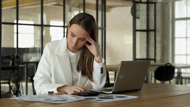 Stylish young female accountant looking through finances, using a laptop and a calculator, experiencing stress due to the fact that the numbers do not add up in the report.