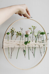 Hand holding stylish spring boho wreath with beautiful fresh flowers. Wooden hoop with flowers and thread. Modern and creative floral handmade decor