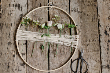 Stylish spring wreath with beautiful fresh flowers. Wooden hoop with flowers and thread, scissors on rustic wooden background flat lay. Modern and creative floral handmade decor