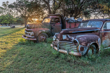 A pair of antique vehicles, a truck and a sedan, rusting away on a grassy field with oak trees and the sun rising in the background, Kendalia, Texas