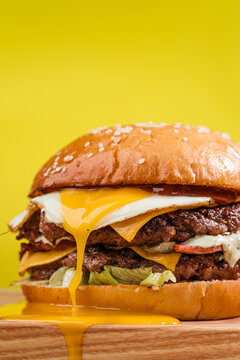 Double patty burger with marbled beef, bacon, cheddar cheese, fried egg with running yolk, tomatoes, onios, cream sauce and ketchup. Wooden table, yellow background. Close up